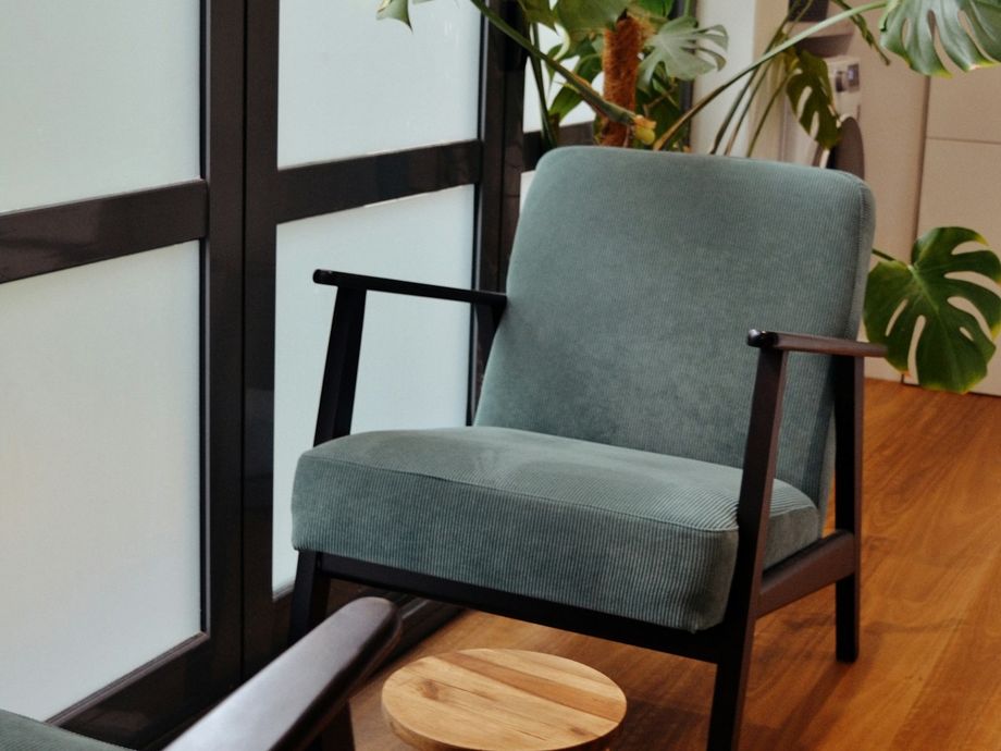 Two green armchairs and a small wooden stool beside a frosted window and a monstera plant in a calm therapy room